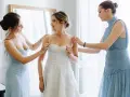 Trois femmes en robes bleues aident la mariée à enfiler sa robe blanche. Une question se pose : à quel âge peut-on être témoin de mariage ?