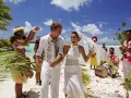 Un jeune couple rayonnant célèbre son mariage à deux à l'étranger sur une plage tropicale, entouré de musiciens et de danseurs locaux.