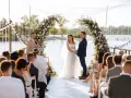 Un couple se tient la main sous une arche fleurie lors d'une cérémonie laïque. Le rituel est célébré au bord de l'eau, devant des invités assis.