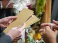 Une main tend une enveloppe dorée contenant des billets, un cadeau de mariage. Une autre main, ornée d'une bague, s'apprête à la recevoir.