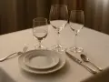 Trois verres à pied sur une table dressée. Quel côté verre à eau est le plus rempli ? Une assiette, une fourchette, un couteau et une cuillère complètent la mise en place.