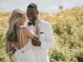 Un couple heureux dans un champ de fleurs jaunes. La mariée porte une robe de mariée blanche, simple et élégante, avec un dos nu. Le marié est en costume blanc.