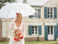 Mariée souriante sous un parapluie blanc, bouquet de fleurs à la main, devant une maison. Un mariage sous la pluie, mais le bonheur est là.