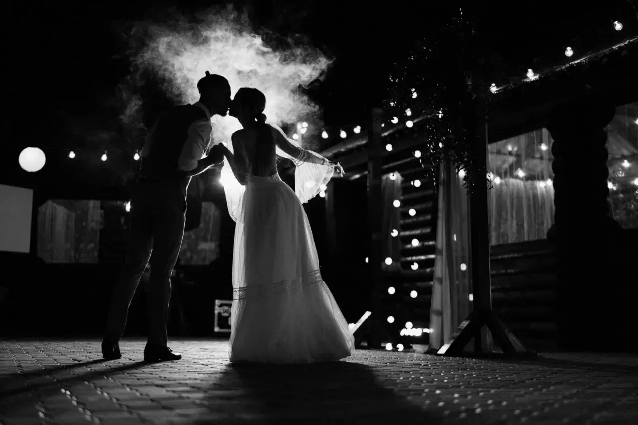 Silhouette d'un couple s'embrassant sous une fumée légère, éclairés par des guirlandes lumineuses. Une scène romantique qui évoque une chanson d'amour.