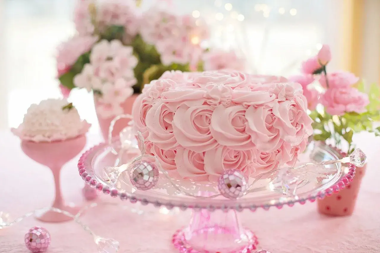 Un délicat **gâteau mariage** rose orné de roses en crème, sur un présentoir scintillant, entouré de fleurs et de lumières féeriques.