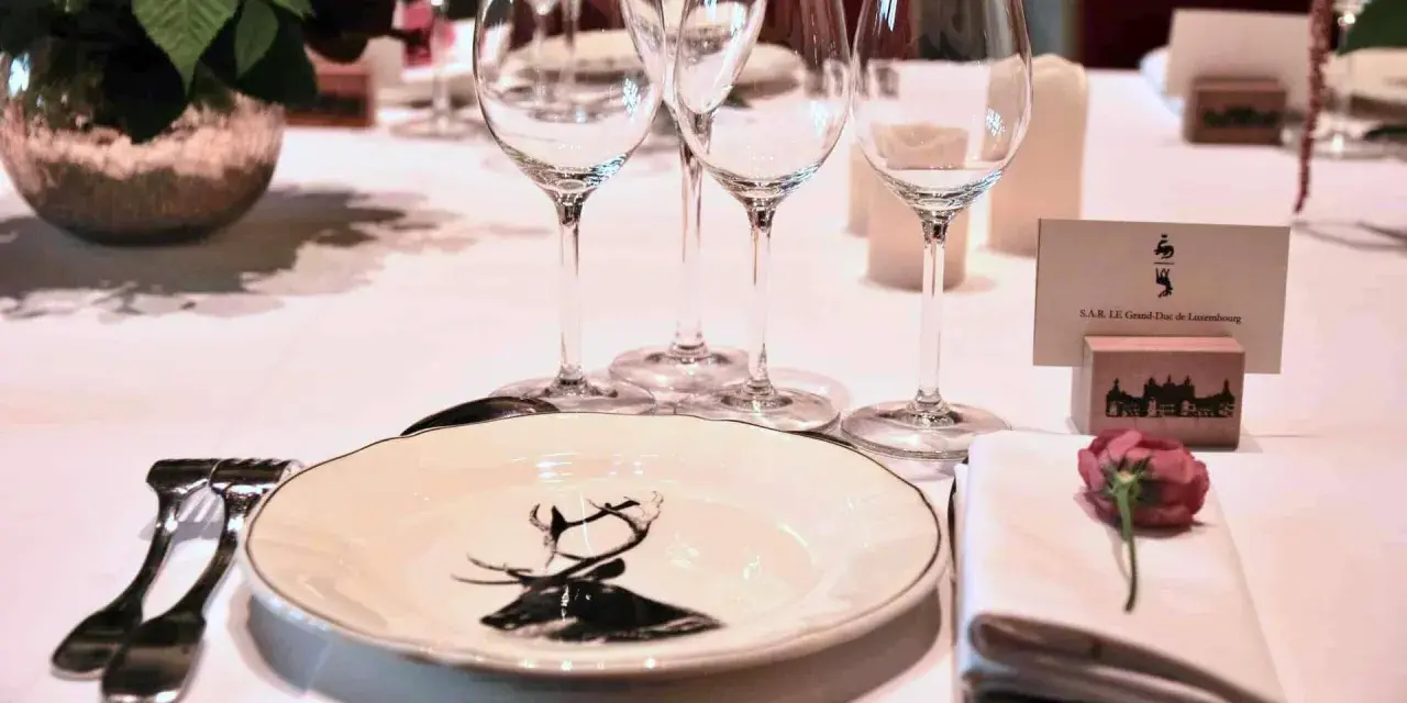 Une élégante table de mariage dressée avec des verres à pied, une assiette ornée d'un cerf, et un marque-place pour le Grand-Duc de Luxembourg.
