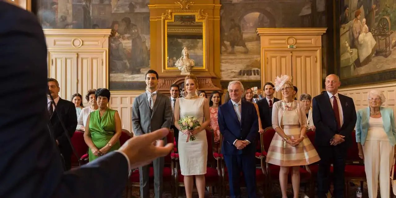 Le couple se tient au centre, entouré de leurs 5 témoins mariage. La mariée porte une robe blanche et tient un bouquet. Les invités sont élégamment vêtus.