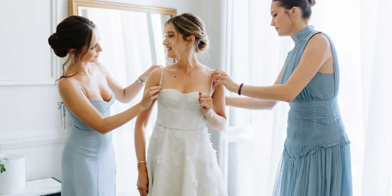 Trois femmes en robes bleues aident la mariée à enfiler sa robe blanche. Une question se pose : à quel âge peut-on être témoin de mariage ?