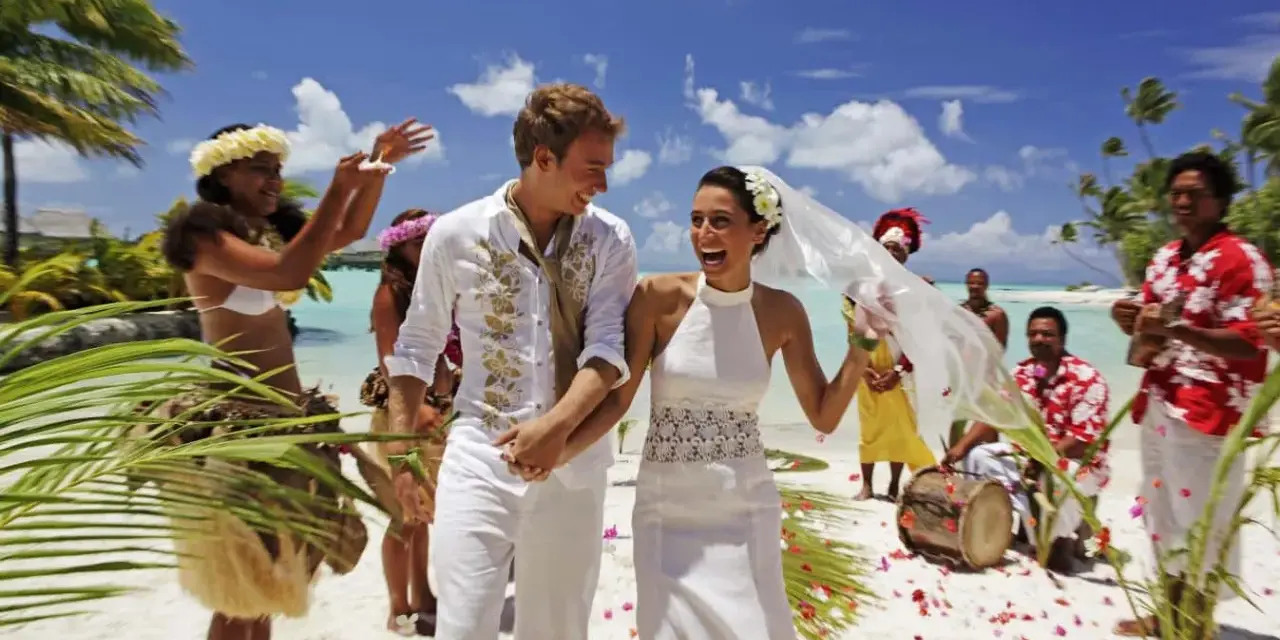 Un jeune couple rayonnant célèbre son mariage à deux à l'étranger sur une plage tropicale, entouré de musiciens et de danseurs locaux.
