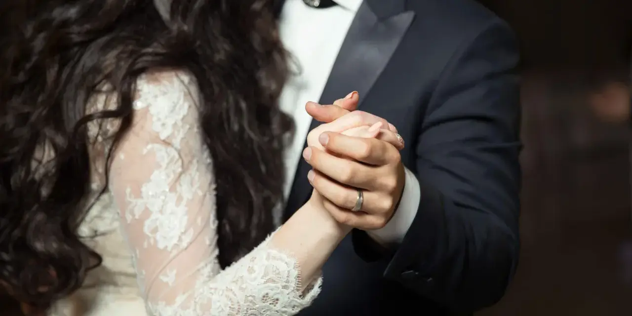 Les mains jointes d'un couple dansant une valse mariage. La mariée porte une robe en dentelle, le marié un costume sombre.