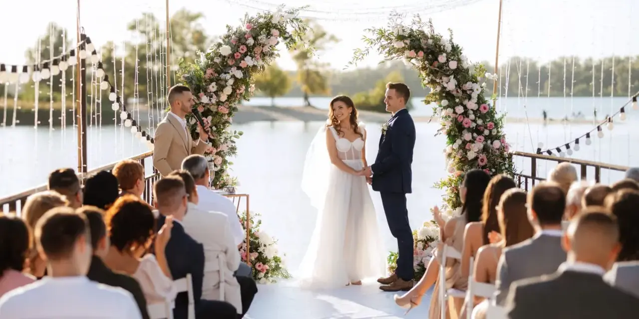 Un couple se tient la main sous une arche fleurie lors d'une cérémonie laïque. Le rituel est célébré au bord de l'eau, devant des invités assis.