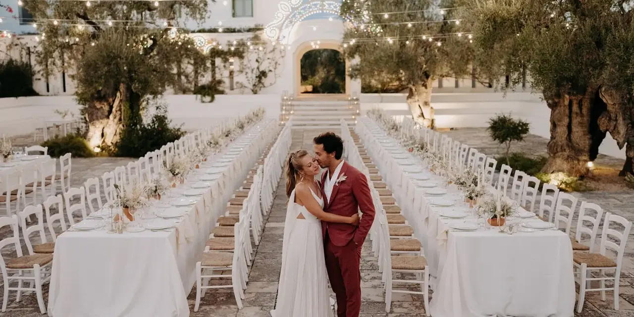 Un couple s'embrasse devant de longues tables dressées pour un mariage dans les Pouilles, illuminé par des guirlandes lumineuses.