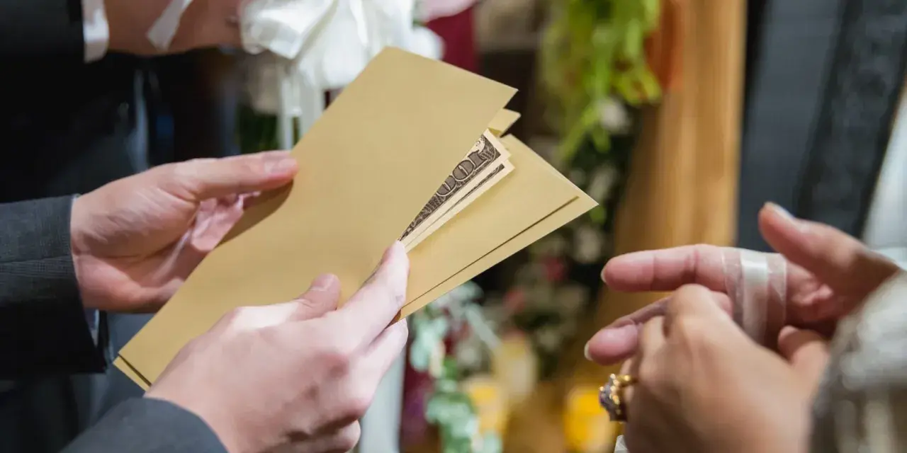 Une main tend une enveloppe dorée contenant des billets, un cadeau de mariage. Une autre main, ornée d'une bague, s'apprête à la recevoir.