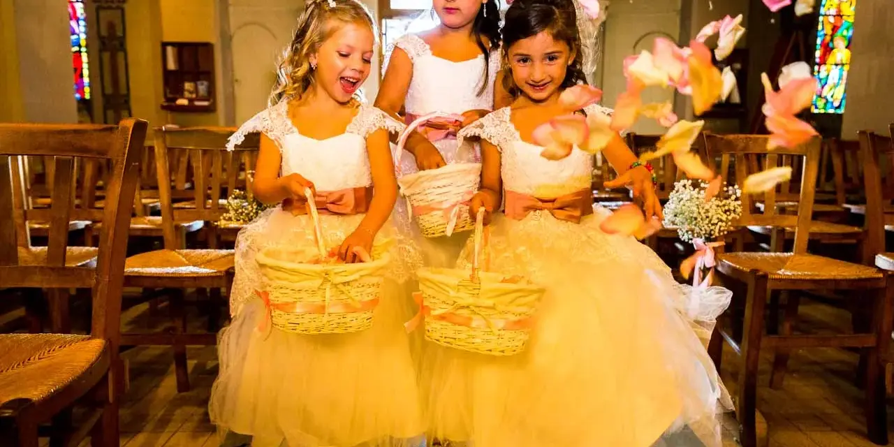 Trois enfants d'honneur souriantes, vêtues de robes claires, tiennent des paniers fleuris. Des pétales de roses volent autour d'elles dans une église.