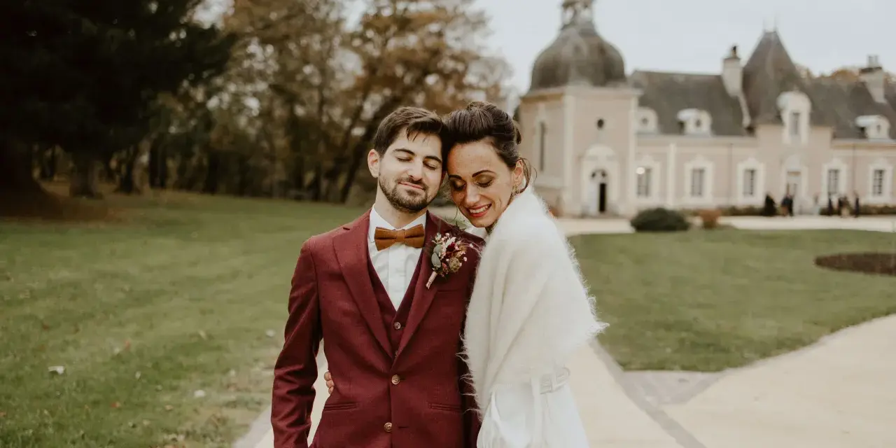 Un jeune marié rayonne dans son costume bordeaux, la couleur costume mariage parfaite pour un jour d'automne. Il enlace tendrement sa mariée devant un château.