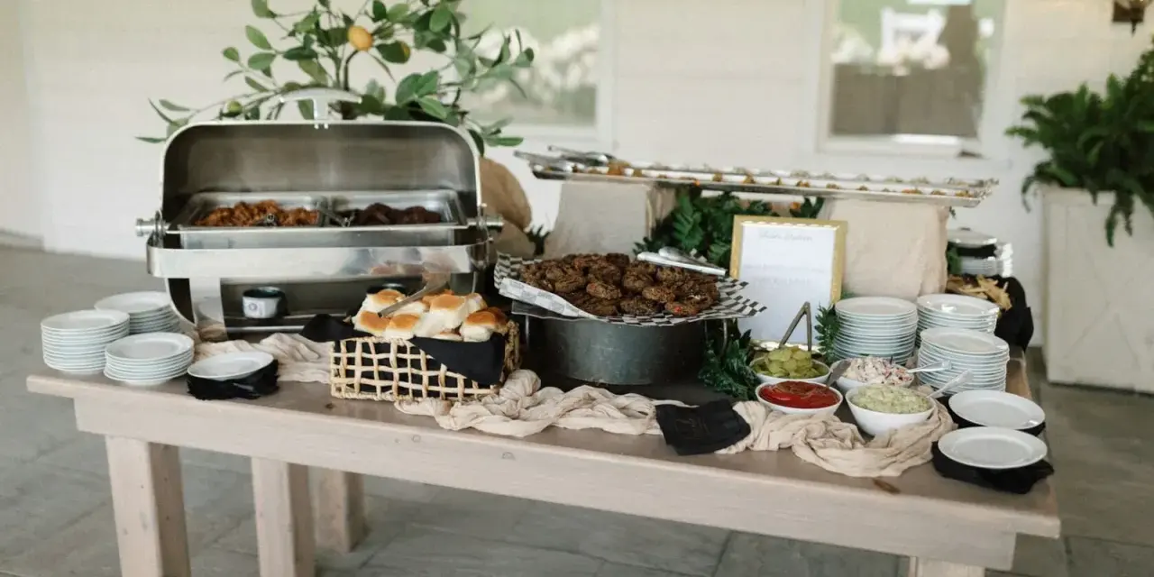 Buffet de brunch mariage avec plats chauds, petits pains, garnitures et assiettes empilées.