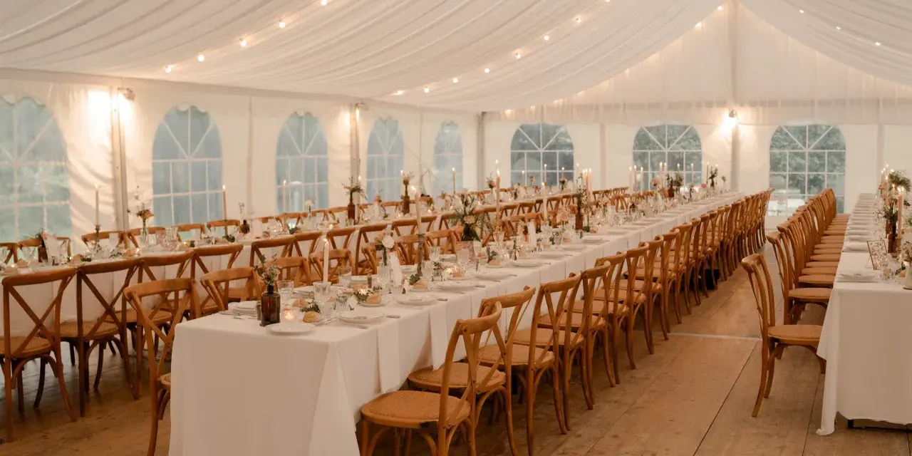 Ambiance chaleureuse sous tente pour un mariage, avec une longue table ronde ou rectangulaire dressée, éclairée par des guirlandes lumineuses et des bougies.