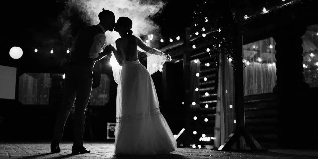 Silhouette d'un couple s'embrassant sous une fumée légère, éclairés par des guirlandes lumineuses. Une scène romantique qui évoque une chanson d'amour.