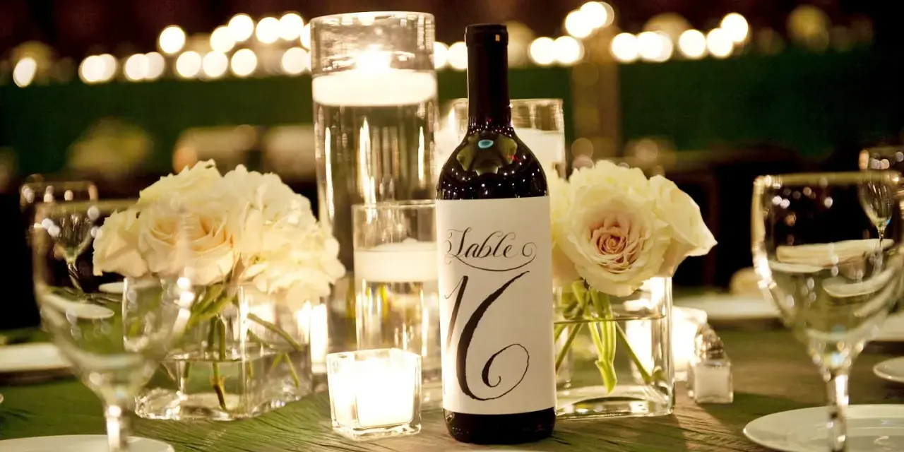 Bouteille de vin avec étiquette "Table 16", entourée de roses pâles, bougies et verres à vin. Parfait pour un dîner avec droit de bouchon traiteur.