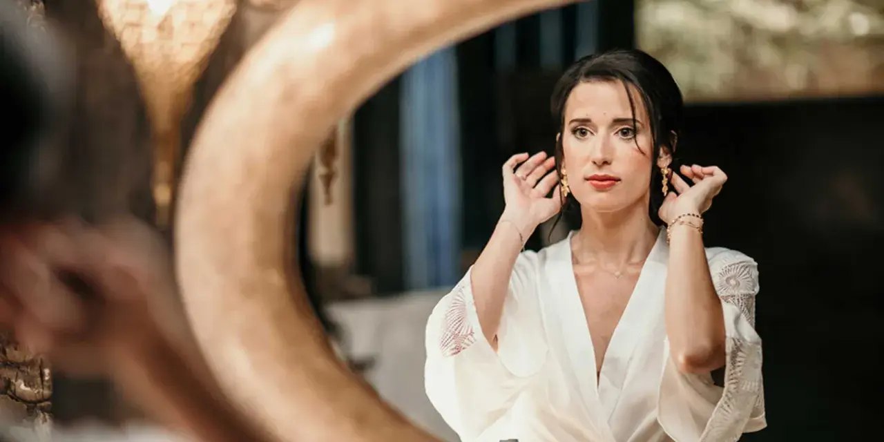 Une mariée, le visage marqué par le stress du mariage, ajuste ses boucles d'oreilles devant un miroir.