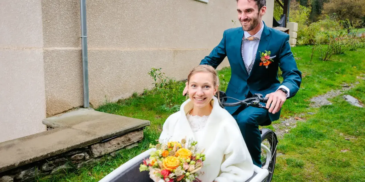 Un jeune couple souriant sur un vélo cargo, la mariée tenant un bouquet. Une idée originale pour un mariage avec un petit budget.