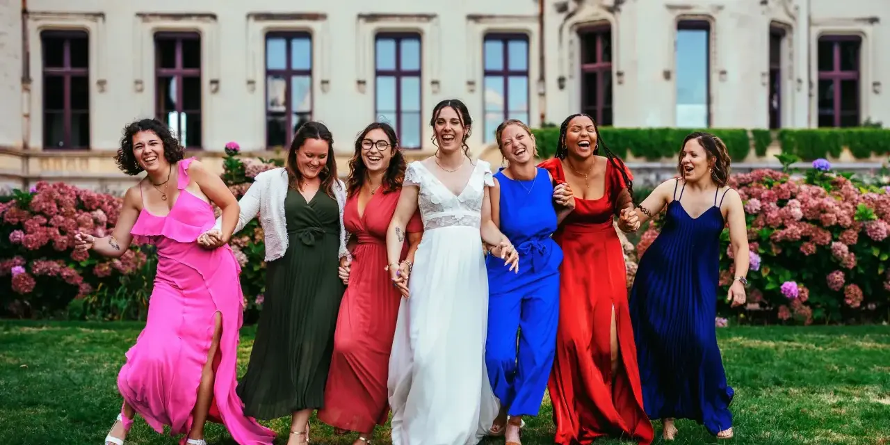 Un groupe de femmes rit joyeusement devant un château. La mariée en blanc est entourée de ses demoiselles d'honneur en robes colorées. La différence entre témoin et demoiselle d'honneur est subtile, mais leur complicité est évidente.