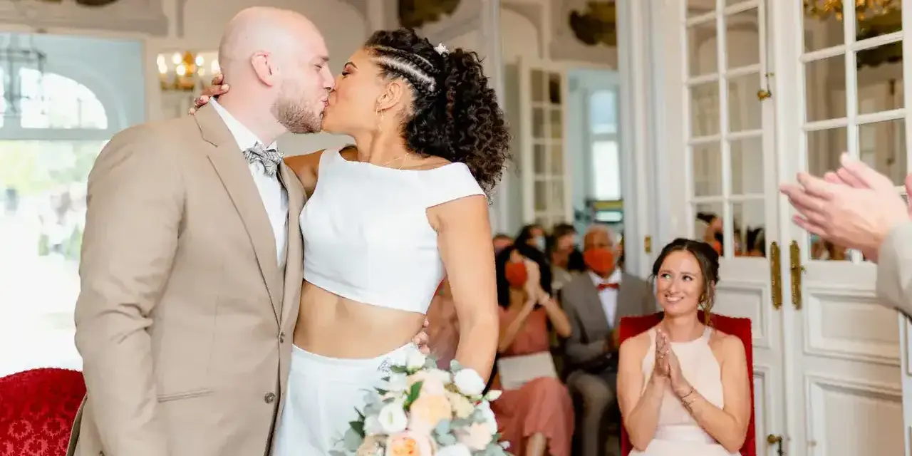 Un couple s'embrasse lors d'un mariage civil sans robe de mariée. La mariée porte un ensemble blanc moderne, le marié un costume beige.