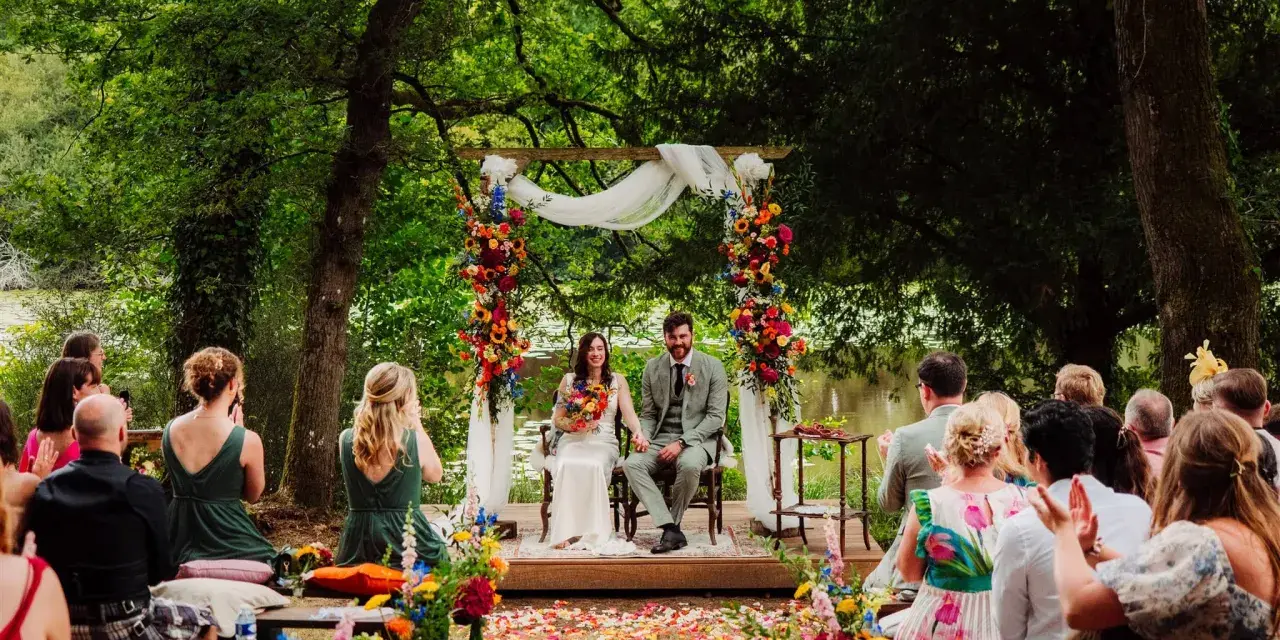 Un couple souriant lors d'une cérémonie laïque en plein air, entouré de verdure et de fleurs colorées. L'officiant cérémonie laïque guide ce moment unique.