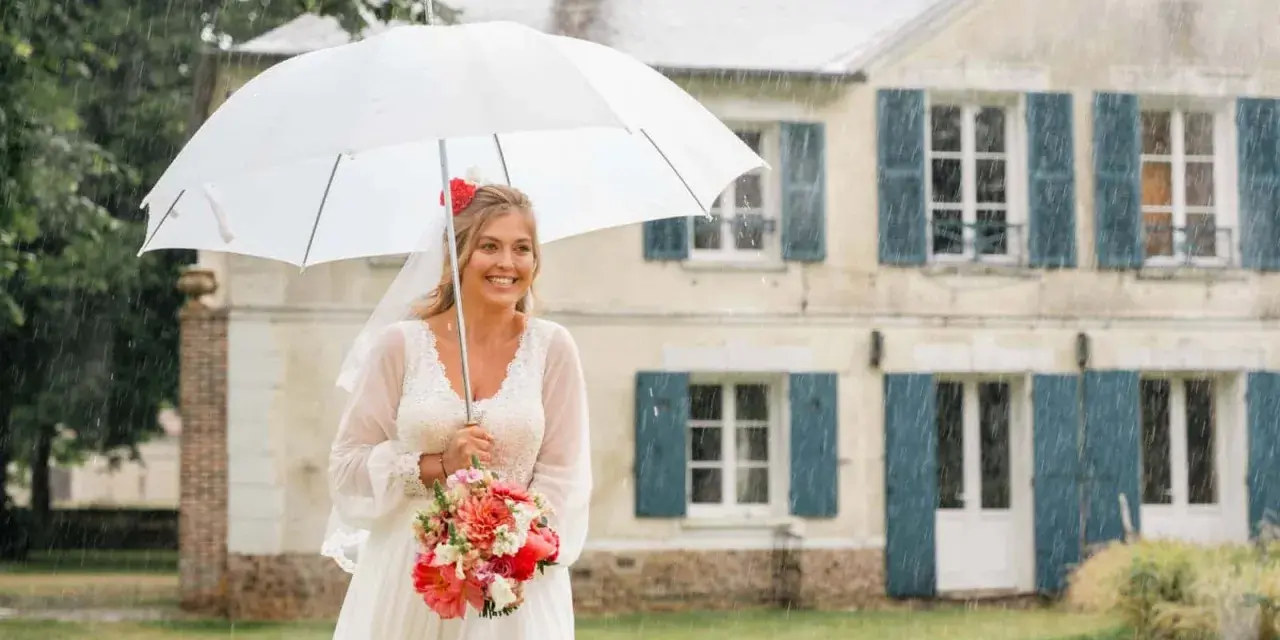 Mariée souriante sous un parapluie blanc, bouquet de fleurs à la main, devant une maison. Un mariage sous la pluie, mais le bonheur est là.