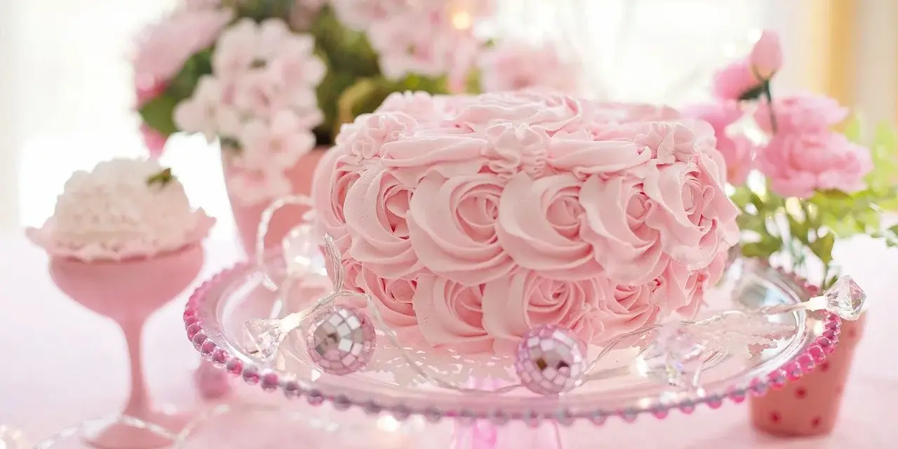 Un délicat **gâteau mariage** rose orné de roses en crème, sur un présentoir scintillant, entouré de fleurs et de lumières féeriques.