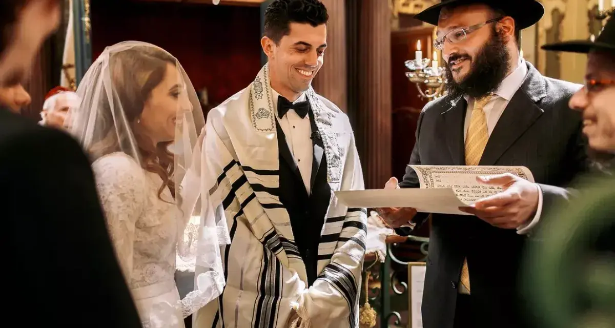 Un couple rayonne lors de leur mariage juif. Le marié, drapé d'un châle de prière, sourit à sa fiancée voilée. Un rabbin lit un document sacré.
