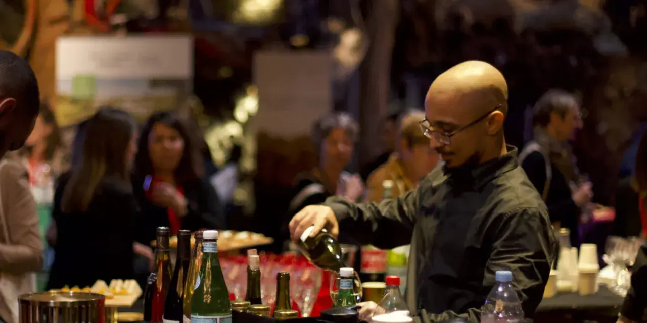 Un barman sert du vin lors d'un vin d'honneur pour 100 personnes. Des bouteilles et des verres sont disposés sur le comptoir.