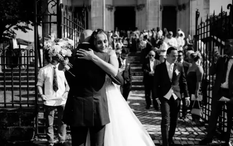 Un couple s'embrasse devant une église, entouré d'invités. Un jeune garçon tient un bouquet. Ce moment est le fruit d'un savant **rétroplanning mariage**.