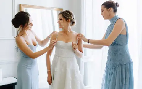 Trois femmes en robes bleues aident la mariée à enfiler sa robe blanche. Une question se pose : à quel âge peut-on être témoin de mariage ?