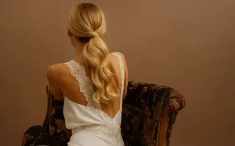 Une mariée aux cheveux blonds, assise sur un fauteuil, arbore une élégante queue de cheval basse mariage, ornée de dentelle sur sa robe.