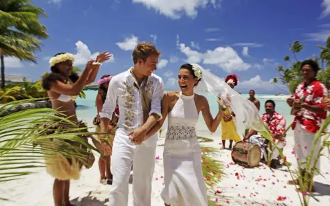 Un jeune couple rayonnant célèbre son mariage à deux à l'étranger sur une plage tropicale, entouré de musiciens et de danseurs locaux.