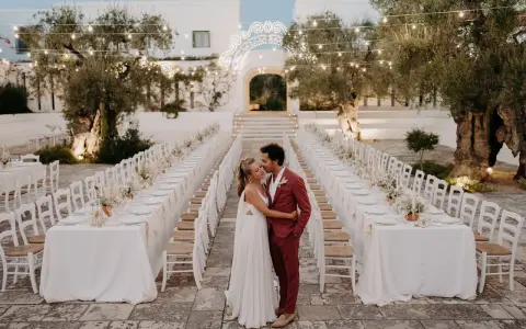 Un couple s'embrasse devant de longues tables dressées pour un mariage dans les Pouilles, illuminé par des guirlandes lumineuses.