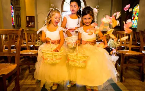 Trois enfants d'honneur souriantes, vêtues de robes claires, tiennent des paniers fleuris. Des pétales de roses volent autour d'elles dans une église.