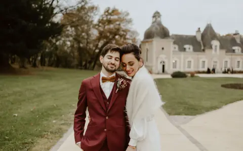 Un jeune marié rayonne dans son costume bordeaux, la couleur costume mariage parfaite pour un jour d'automne. Il enlace tendrement sa mariée devant un château.