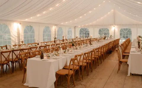 Ambiance chaleureuse sous tente pour un mariage, avec une longue table ronde ou rectangulaire dressée, éclairée par des guirlandes lumineuses et des bougies.