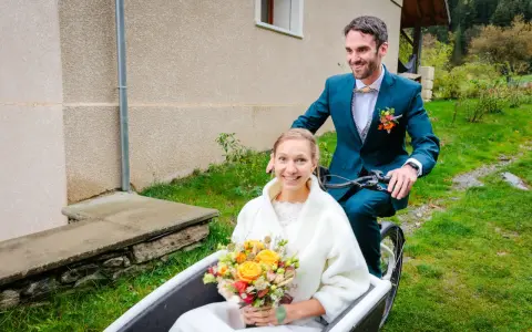 Un jeune couple souriant sur un vélo cargo, la mariée tenant un bouquet. Une idée originale pour un mariage avec un petit budget.