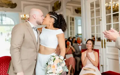 Un couple s'embrasse lors d'un mariage civil sans robe de mariée. La mariée porte un ensemble blanc moderne, le marié un costume beige.