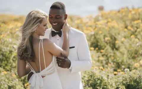 Un couple heureux dans un champ de fleurs jaunes. La mariée porte une robe de mariée blanche, simple et élégante, avec un dos nu. Le marié est en costume blanc.