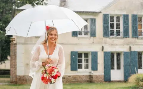 Mariée souriante sous un parapluie blanc, bouquet de fleurs à la main, devant une maison. Un mariage sous la pluie, mais le bonheur est là.