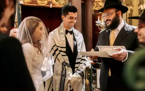 Un couple rayonne lors de leur mariage juif. Le marié, drapé d'un châle de prière, sourit à sa fiancée voilée. Un rabbin lit un document sacré.