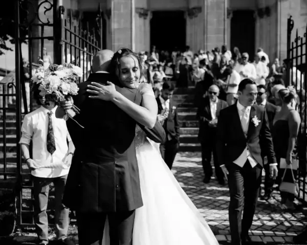Un couple s'embrasse devant une église, entouré d'invités. Un jeune garçon tient un bouquet. Ce moment est le fruit d'un savant **rétroplanning mariage**.