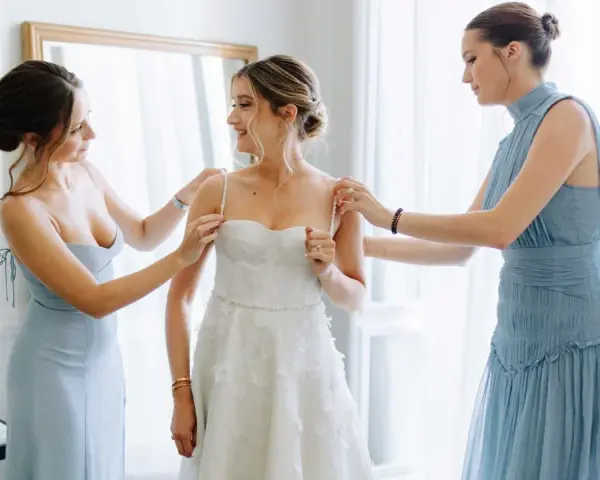 Trois femmes en robes bleues aident la mariée à enfiler sa robe blanche. Une question se pose : à quel âge peut-on être témoin de mariage ?