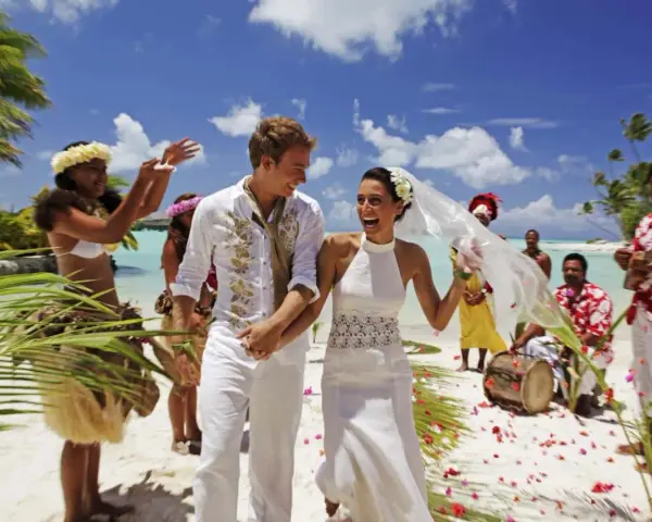 Un jeune couple rayonnant célèbre son mariage à deux à l'étranger sur une plage tropicale, entouré de musiciens et de danseurs locaux.