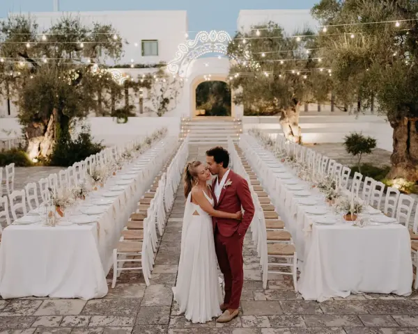 Un couple s'embrasse devant de longues tables dressées pour un mariage dans les Pouilles, illuminé par des guirlandes lumineuses.