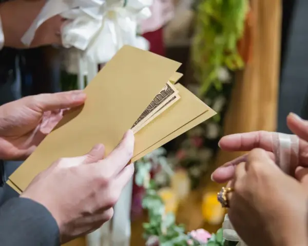 Une main tend une enveloppe dorée contenant des billets, un cadeau de mariage. Une autre main, ornée d'une bague, s'apprête à la recevoir.
